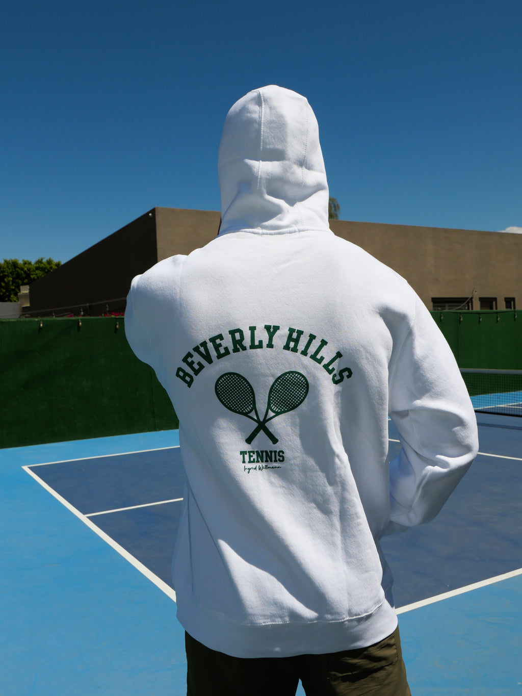 Person wearing a white hoodie with 'Beverly Hills Tennis' text and graphics on a tennis court.